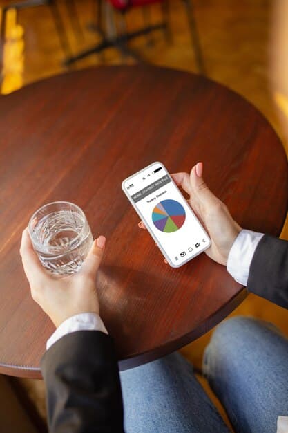 A person using a budgeting app on their smartphone while sitting at a coffee shop. The app displays a colorful pie chart showing categorized expenses.